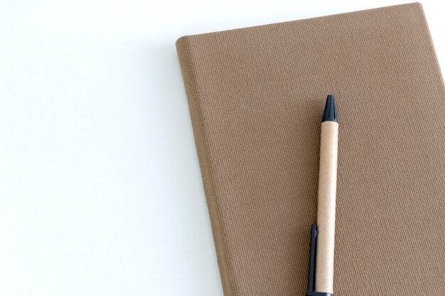 Top view of brown notebook and a pen on white desk background