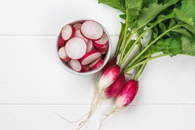 Top view of a bunch of radishes and a bowl of radish slices on a white wooden table. A fresh crop of radishes.