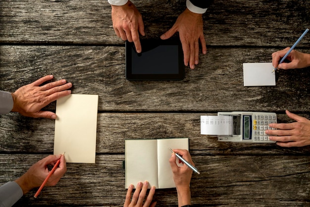 Photo top view of businessmen hands with holding notes, tablet computer and printing calculator on top of a rustic wooden table.