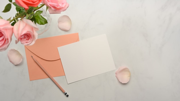 Top view of card, pastel envelope, pencil and pink roses flower decorated on marble desk