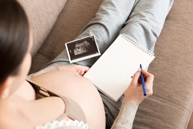 Photo top view caring future mother writing diary with ultrasound pregnant woman tummy making notes feeling during pregnancy or creating scrapbook