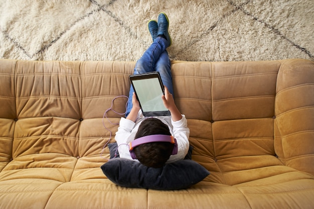 Вид сверху кавказского мальчика в шлемах, играющего с планшетом, сидя на диване у себя дома