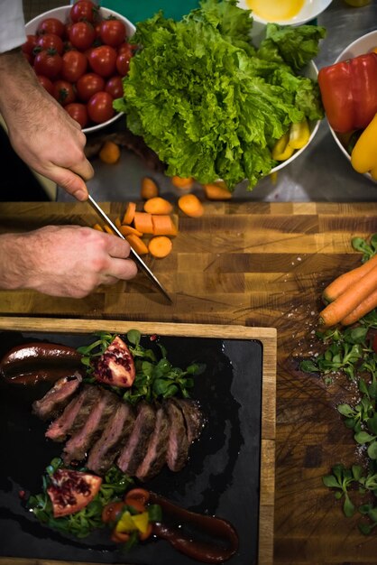 Вид сверху на руки шеф-повара на кухне отеля или ресторана, где подают стейк из говядины с овощным украшением