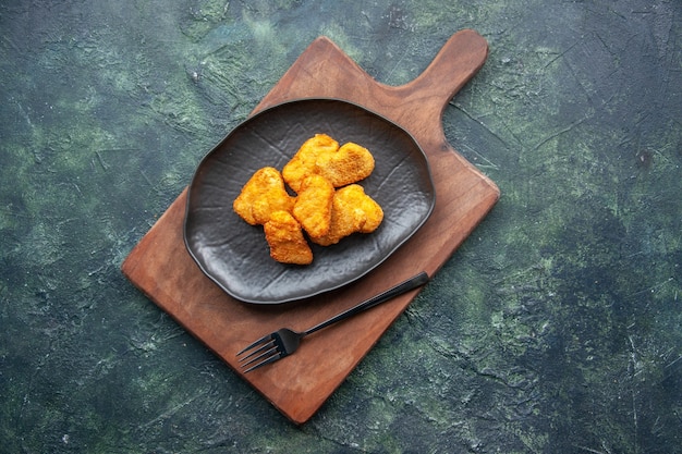 Vista dall'alto di pepite di pollo su un piatto nero e forchetta sul tagliere di legno sulla superficie scura