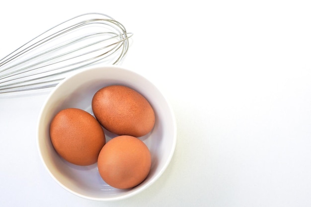 Top view of chickken eggs in a bowl and egg whisks on white background Copy space