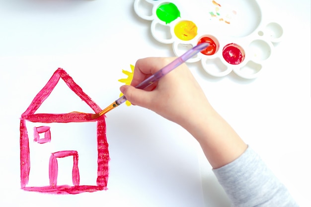 Top view of child hand with pencil is drawing picture of red house on white paper.