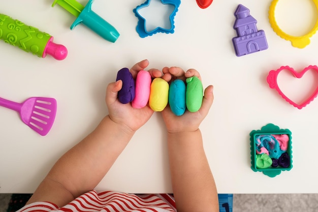 Top view children's hands with multicolored plasticine or playdough on white table with toys children's creativity