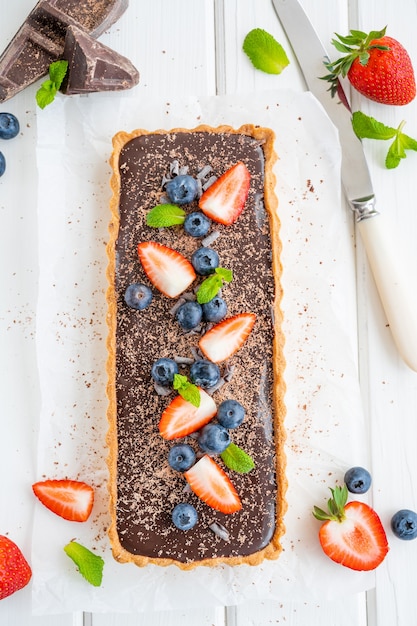 Top view chocolate cream tart with fresh blueberries and strawberries on white wooden background