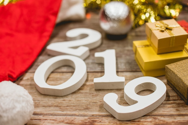 Photo top view of christmas hat, 2019 number and gift boxes on wooden background.