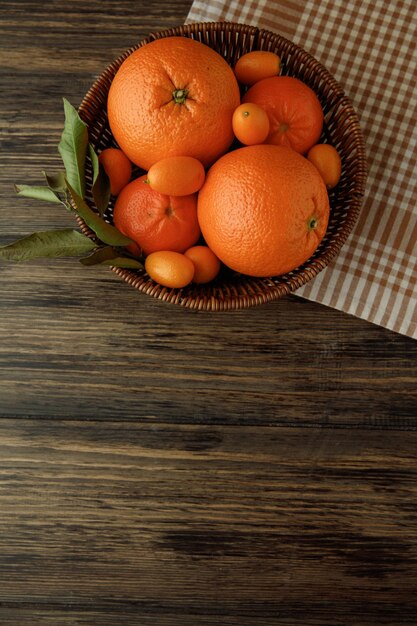 Top view of citrus fruits as orange tangerine kumquat in basket on plaid cloth on wooden background with copy space