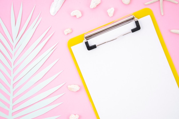 Top view clipboard with a blank piece of paper and palm leaf on pink background