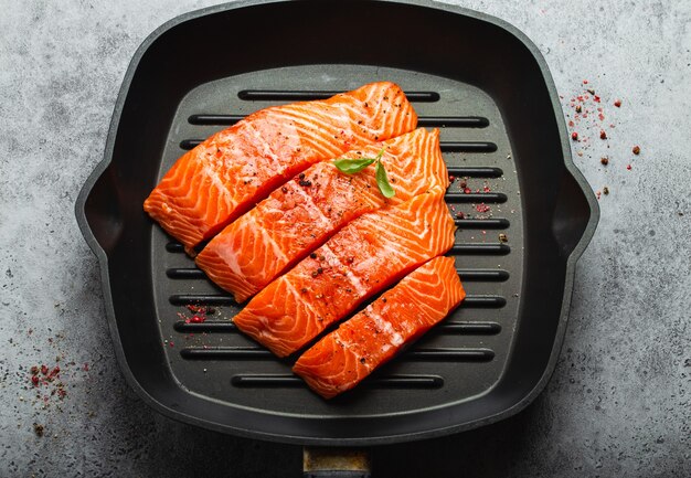 Top view, close-up of cut in slices fresh raw salmon fillet garnished with green basil leaves on grill skillet, gray stone background. Preparing salmon fillet for cooking, healthy eating concept