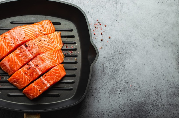Top view, close-up of cut in slices fresh raw salmon fillet with seasonings on grill skillet, gray stone background. Preparing salmon fillet for cooking, healthy eating concept. Space for text