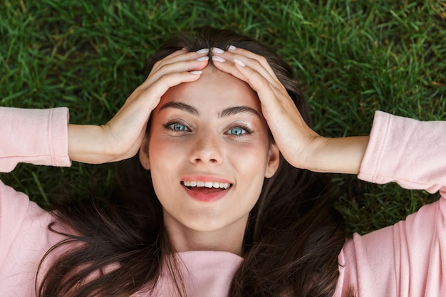 Top view close up of a lovely young brunette woman laying on grass outdoors, laughing