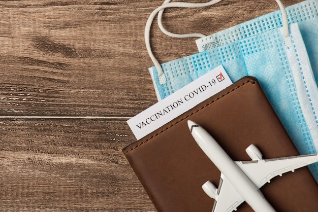 Top view closeup photo of plane model on leather passport cover with vaccination certificate and medical masks on isolated wooden background