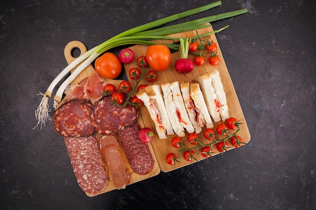 Top view of club sandwiches next to a board with appetizers on dark wooden table