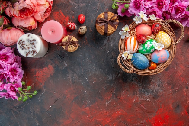 top view colored easter eggs inside basket with flowers and biscuits on dark surface