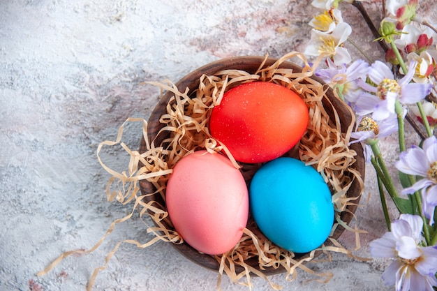 top view colored easter eggs inside plate with straw and flowers on white surface