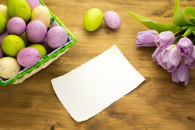 Photo top view of a colorful easter eggs in basket, purple tulips and message card on brown wood background.