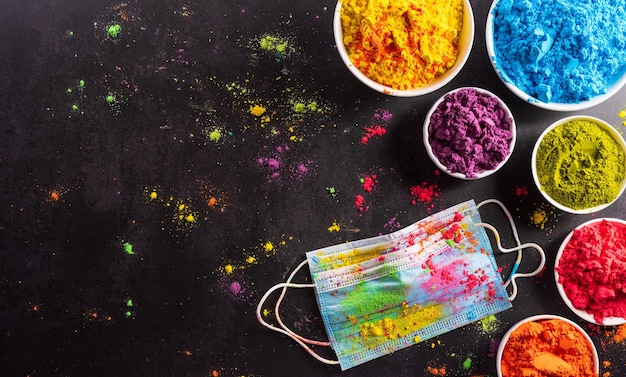 Photo top view of colorful holi powder with multicolored medical mask holi paint on dark background happy holi festival during coronavirus xa covid19 pandemic concept