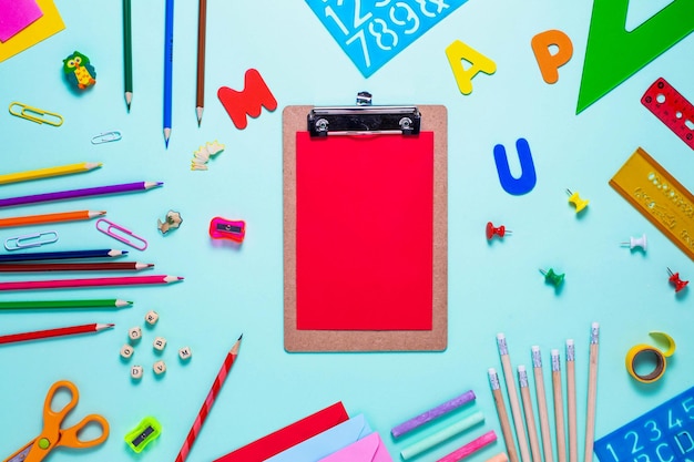 Top view of colorful school supplies with clipboard with red sheet of paper on blue background.