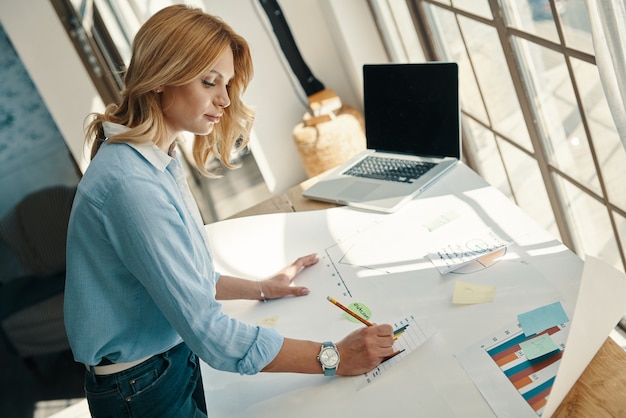 Top view of confident young woman sketching on the blueprint while working in office