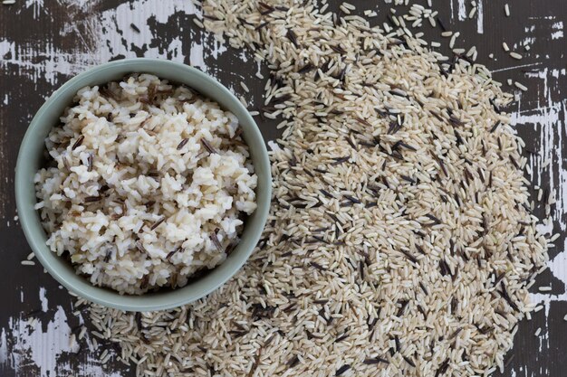 Top view cooked mixed brown and black rice in bowl and heap of raw mixed brown and black rice on the wooden background