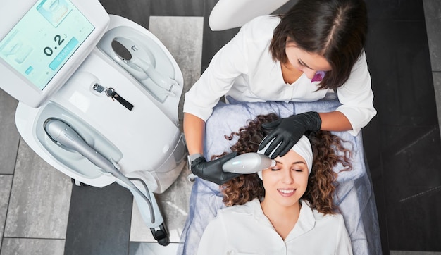 Foto vista dall'alto di un cosmetologo che fa l'hydro peeling al vuoto sulla fronte per una giovane paziente donna sorridente concetto di procedura di processo con attrezzature moderne in una clinica professionale