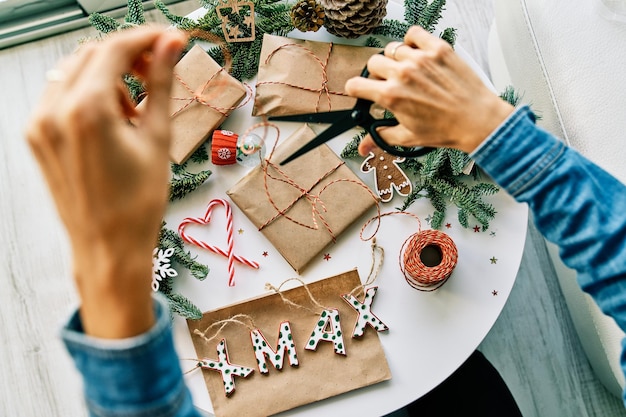 Top view of crop anonymous female with scissors cutting ribbon while preparing Christmas presents on table with decorations