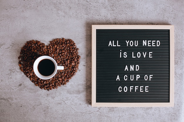 Вид сверху на чашку кофе на красивой форме сердца из кофейных зерен с цитатой на доске для писем говорит, что все, что вам нужно, это любовь и чашка кофе