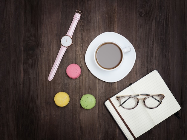 一杯のコーヒー、マカロン、ノート、時計、メガネの平面図です。木製の背景