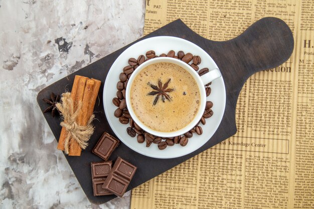 top view cup of coffee with anise biscuits roasted coffee beans on saucer chocolate cinnamon sticks on wood board newspaper on table