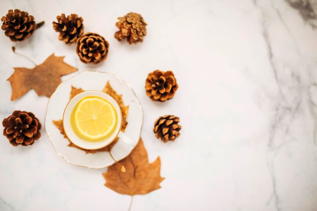 Top view of a cup of lemon tea surrounded pine cones and autumnal maple leaves focus on lemon