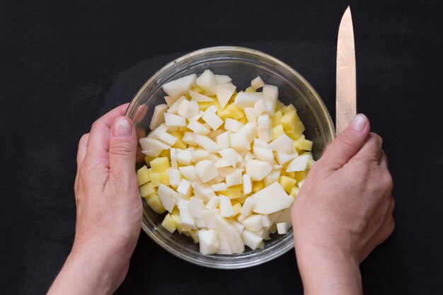 Top view of cut raw potato in glass bowl and woman hands with knife on the black background