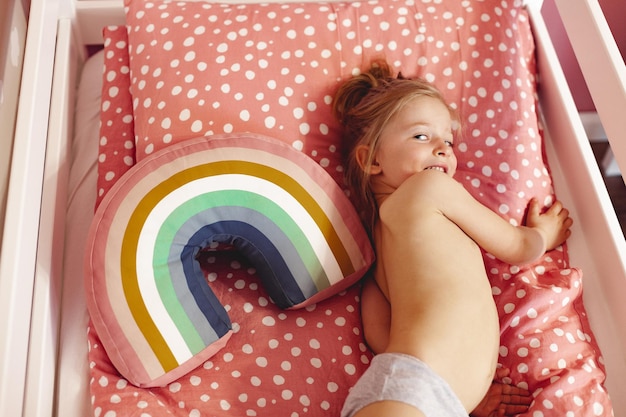 Вид сверху милой просыпающейся маленькой девочки в постели с радужной подушкой