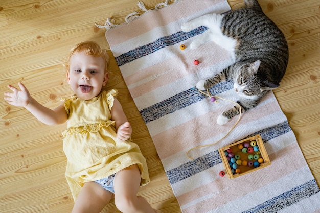 Photo top view cute baby girl in dress having fun lying on floor with cat playing wooden removable beads