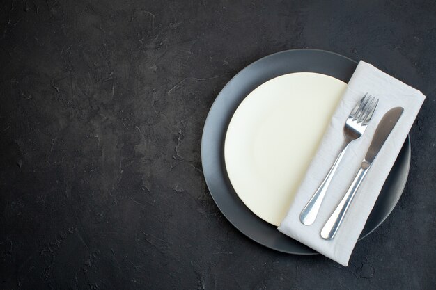 Top view of cutlery set on white napkin on dark color and white empty plates in different sizes on black background