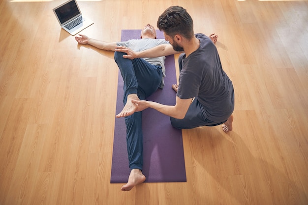 Top view of a dark-haired young yoga instructor stretching his client on the exercise mat