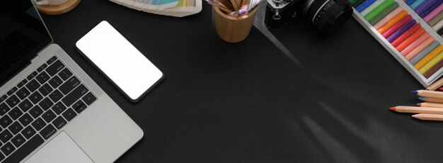 Top view of dark modern worktable with smartphone, laptop, designer supplies and copy space