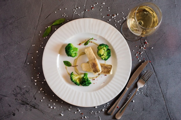 Top view of the delicious lunch with white fish and broccoli on the plate and a glass of wine or juice.