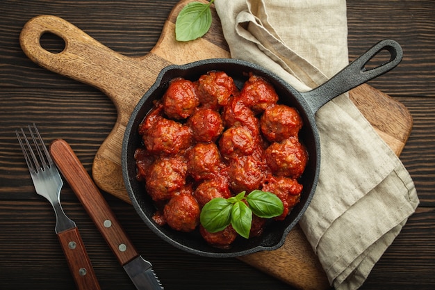 Top view of delicious meatballs with tomato sauce and fresh basil in cast iron rustic vintage pan served on cutting board, wooden background. Tasty homemade meatballs