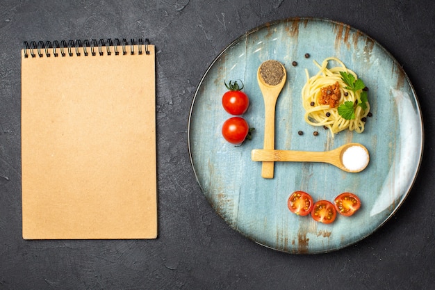 Top view of delicious pasta meal served with meat greens tomatoes on a plate and cutlery set spiral notebook on black background