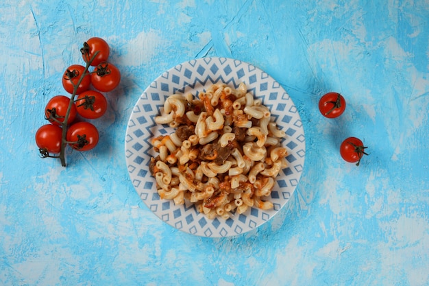 Top view of delicious pasta with meat pieces and sauce on the beautiful plate on the blue textured table, fresh tomatoes decorate the serving. Copy space.