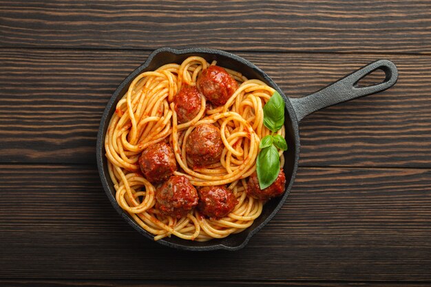 Top view of delicious pasta with meatballs, tomato sauce and fresh basil in cast iron rustic vintage pan served on cutting board, wooden background. Tasty homemade meatballs spaghetti