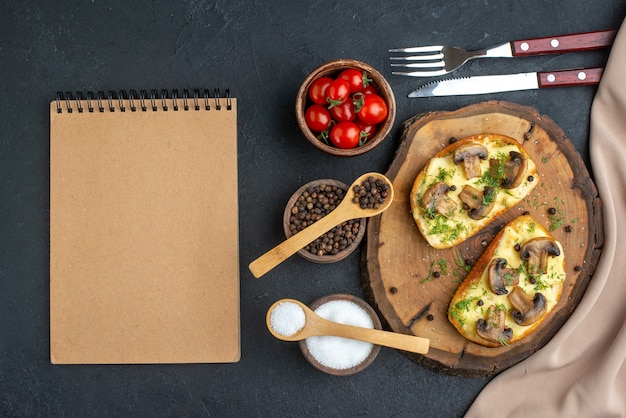 Вид сверху вкусной закуски с грибами на деревянной доске и набор столовых приборов, перец, помидоры, ноутбук на черном фоне