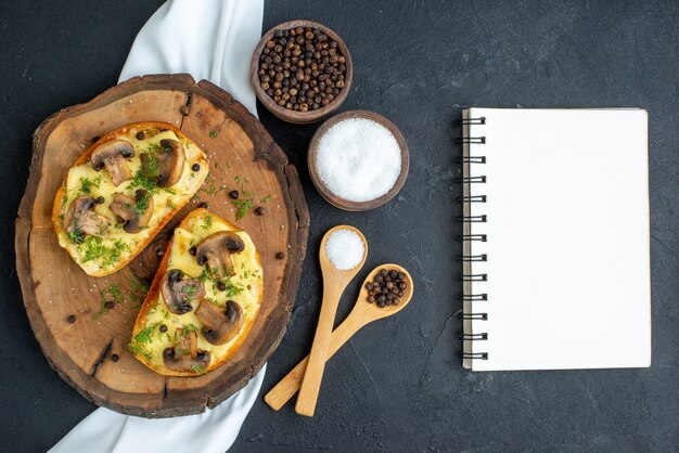 Top view of delicious snack with mushrooms on wooden board and spices on white towel notebook on black background