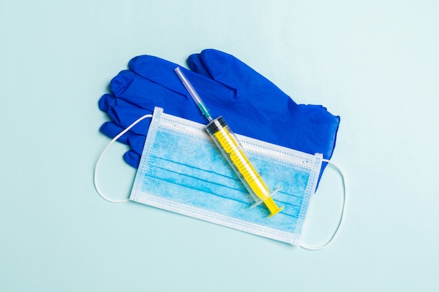 Top view of disposable surgical mask, pair of latex medical gloves and syringe on blue surface