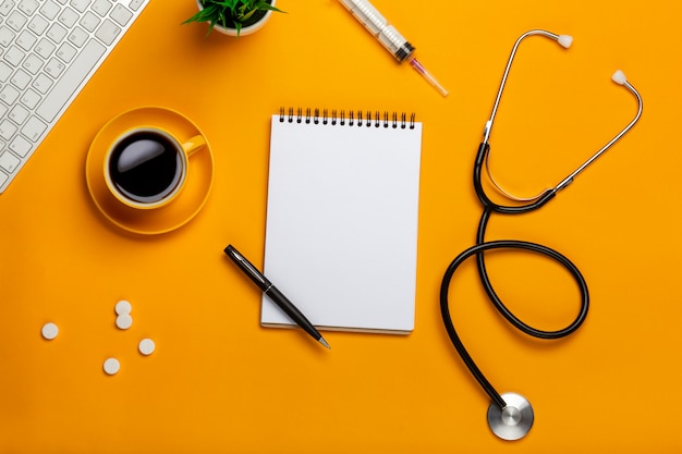 Vista dall'alto del tavolo di un medico con blocco note e penna stetoscopio, tastiera, prescrizione e pillole, una tazza di caffè su uno sfondo giallo