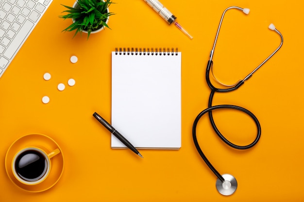 Top view of a doctor's table with notepad and pen stethoscope, keyboard, prescription and pills, a cup of coffee on a yellow background