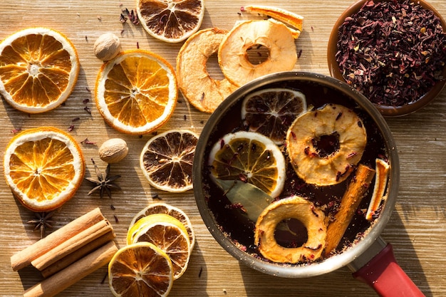 Top view of dry pieces of lemon, orange and apple, cinnamon sticks, bay leaves, nutmegs, star anise and dry cutted hibiscus and the saucepan with water and ingredients in it